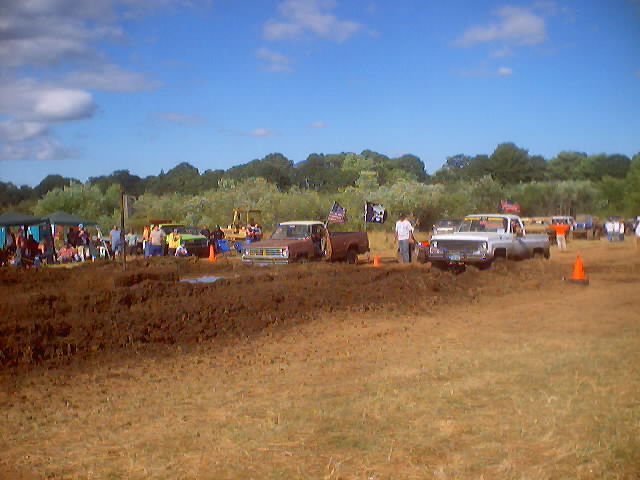 Mud bogging event with several trucks in the mud, spectators under tents, and trees in the background