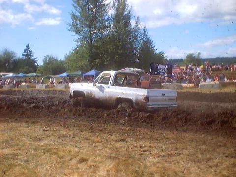 1976 Ford F-100 - A white pickup truck navigates a muddy track during a mud drag event, with spectators in the background.