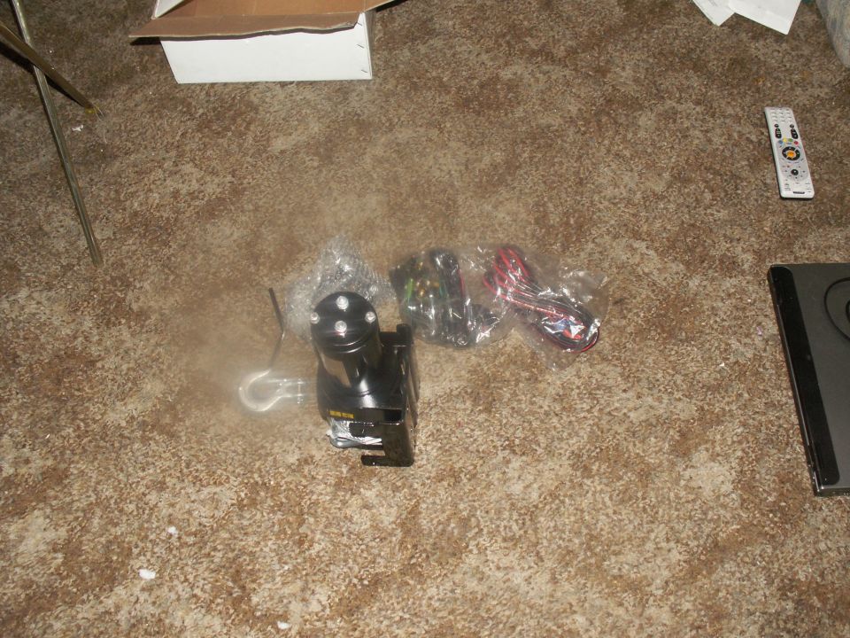Black winch with hook and wiring, remote control, and box on a carpeted floor.