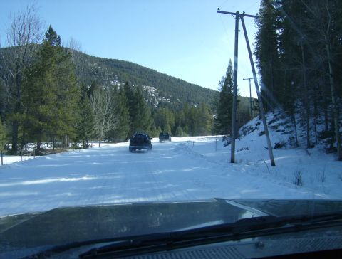 1990 Ford Bronco - A dark colored SUV drives down a snow-covered road in a mountainous, forested area. The view is from the interior of another vehicle, looki…
