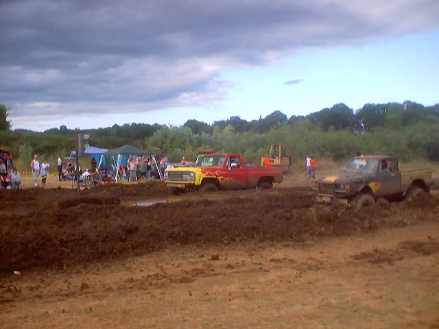 Two trucks stuck in mud at an outdoor event with spectators and tents in the background