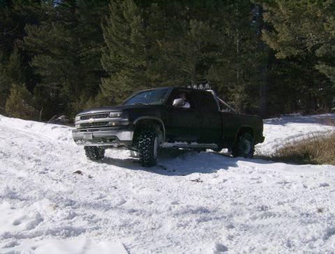 1990 Ford Bronco - A black pickup truck with off-road tires is parked on a snowy incline in front of a dense forest. The truck has a roll bar with auxiliary l…