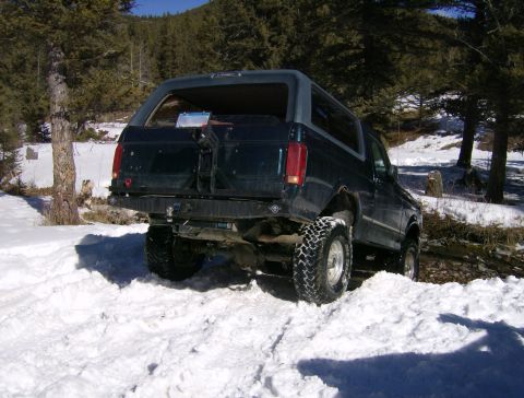 1990 Ford Bronco - The rear passenger side of a dark blue Ford Bronco is shown parked in deep snow, with a forest and mountains in the background. The vehicle…