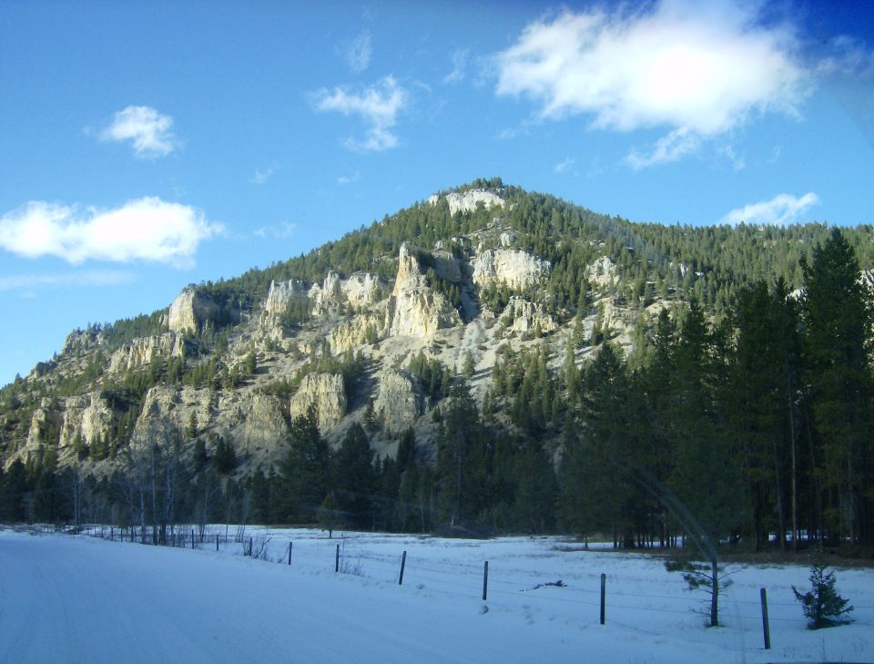 Snowy road leading to a forested mountain range with rocky cliffs.