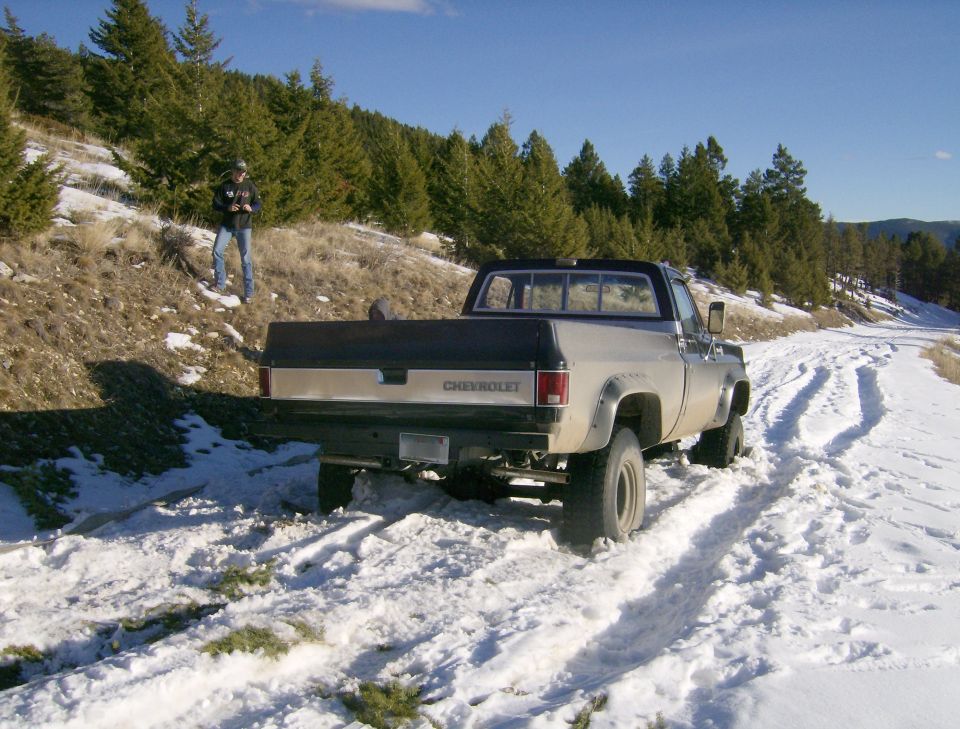Silver and black Chevrolet pickup truck on a snowy road with a person on a hillside.