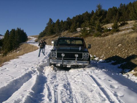 1990 Ford Bronco - A black pickup truck is stuck in the snow on a dirt road, with two people standing nearby. The truck has a chrome bumper and dual headlight…