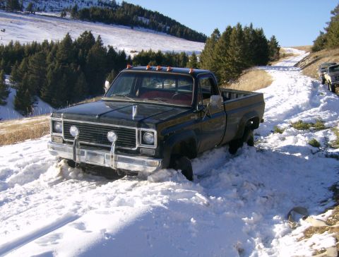 1990 Ford Bronco - A dark-colored pickup truck is stuck in deep snow on a dirt road in a wooded, hilly area. The truck is facing towards the viewer with its f…