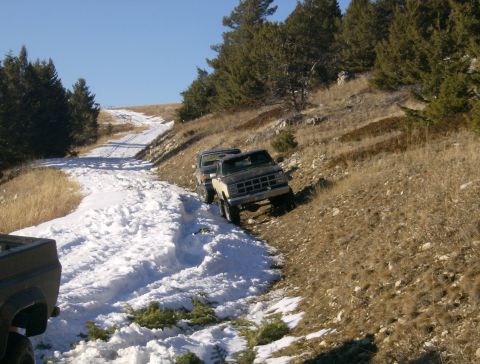 1990 Ford Bronco - Two pickup trucks navigate a snow-covered dirt road on a hillside with scattered pine trees under a clear blue sky.