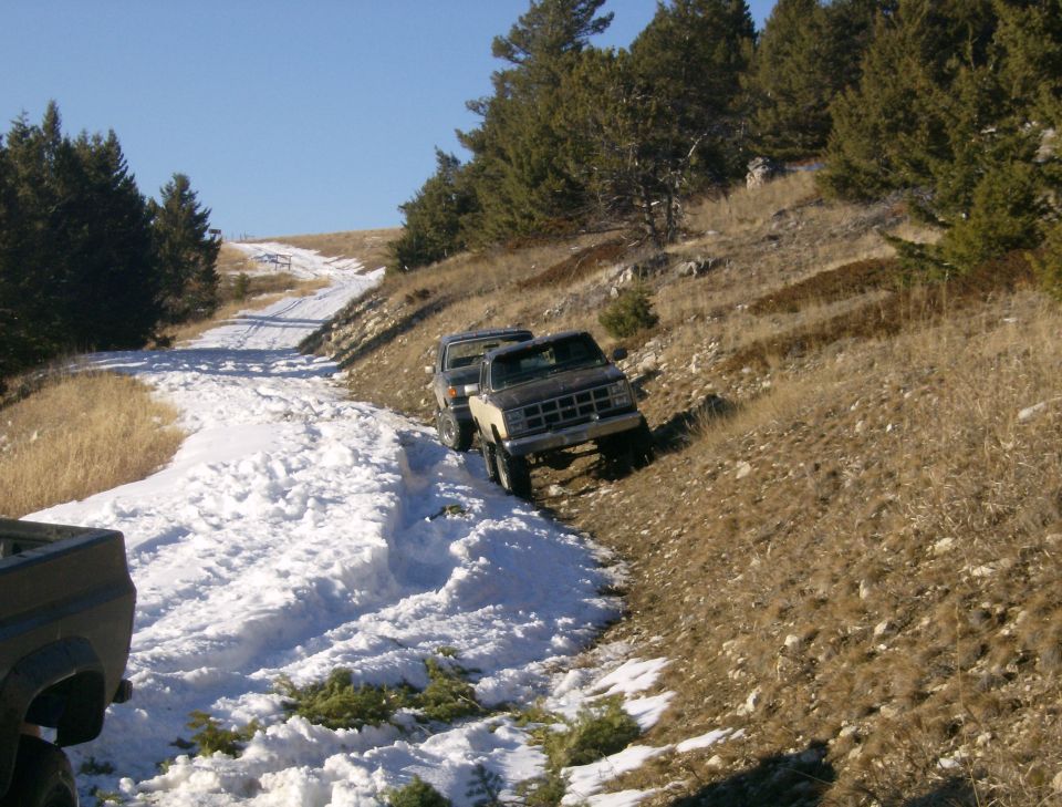 Two pickup trucks on a snowy, hilly road with pine trees.