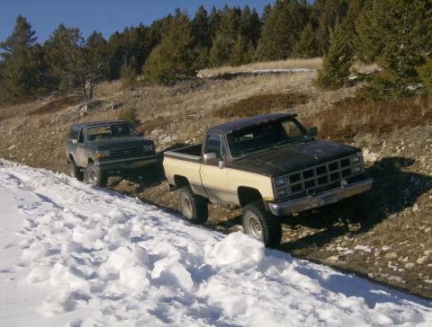 1990 Ford Bronco - Two off-road vehicles, a dark green SUV and a two-tone pickup truck, are parked on a snowy, uneven dirt road with trees in the background.