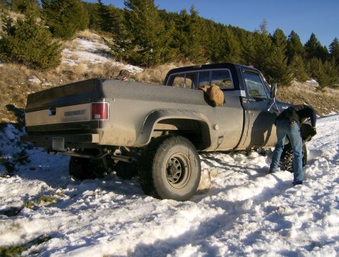 1990 Ford Bronco - A person shovels snow from around the rear tire of a dusty, dark gray Chevrolet pickup truck parked on a snowy, wooded hillside.