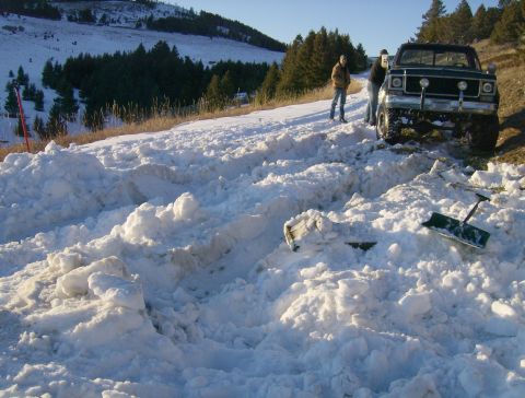 1990 Ford Bronco - A dark green pickup truck is stuck in deep snow on a rural road, with two people standing nearby and shovels on the ground.