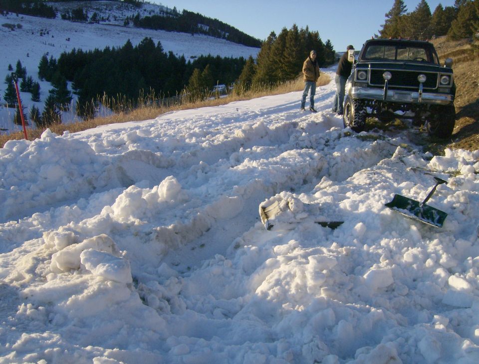 Dark green pickup truck stuck in snow with two people and shovels.