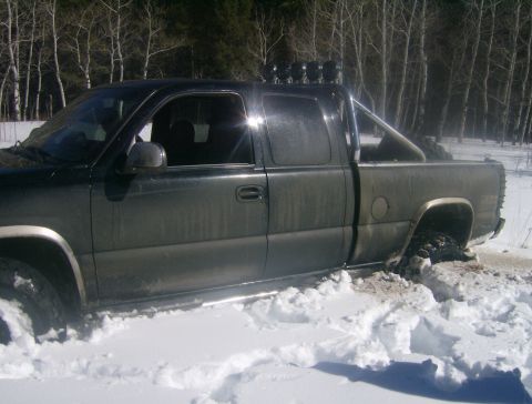 1990 Ford Bronco - A dark-colored pickup truck is stuck in deep snow, with its tires partially submerged. The truck has a roll bar with lights mounted above t…
