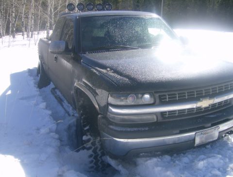 1990 Ford Bronco - A dark-colored pickup truck is stuck in deep snow, with its tires partially submerged. The truck has four auxiliary lights mounted on the r…
