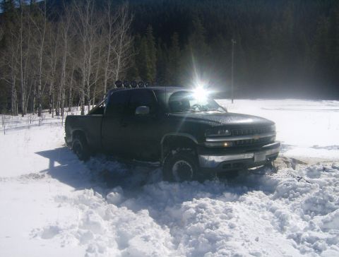 1990 Ford Bronco - Pickup truck stuck in snow with trees and mountains in background. Supermotors.net logo present.