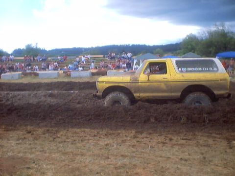1976 Ford F-100 - A yellow Ford Bronco navigates a muddy track during a competition, with a crowd watching from bleachers.