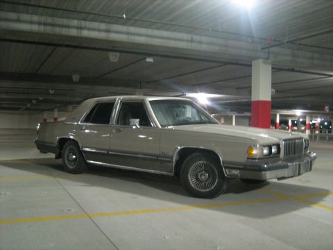 1988 Mercury Grand Marquis - A tan 1988 Mercury Grand Marquis sedan is viewed from the front three-quarter angle in a dimly lit parking garage.