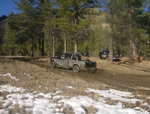 1990 Ford Bronco - Two trucks navigate a challenging off-road course in a forest setting.