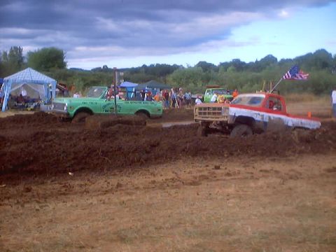 1976 Ford F-100 - Two trucks are stuck in the mud at a drag event, one with an American flag flying from its antenna.