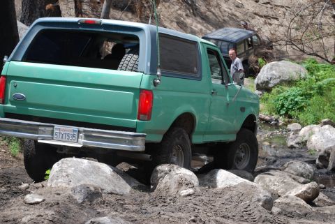 1996 Ford Bronco - A green Ford Bronco is shown off-roading on a rocky trail, with another vehicle visible in the background.