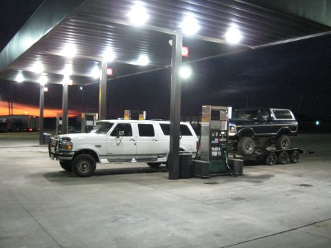 1978 Ford Bronco - A dark-colored Ford Bronco is loaded onto a trailer at a gas station, with a white SUV parked nearby.