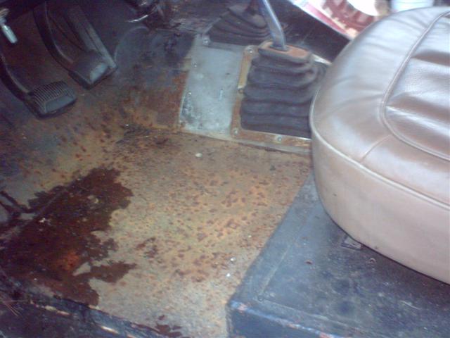 rusted floorboards, pedals, and part of a tan seat in a vehicle interior
