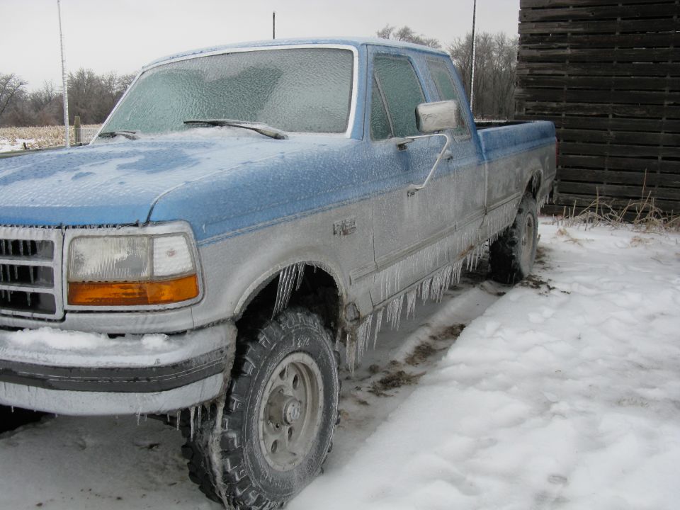 Front 3/4 view of a blue and silver pickup truck covered in ice and icicles, parked in the snow next to a wooden structure