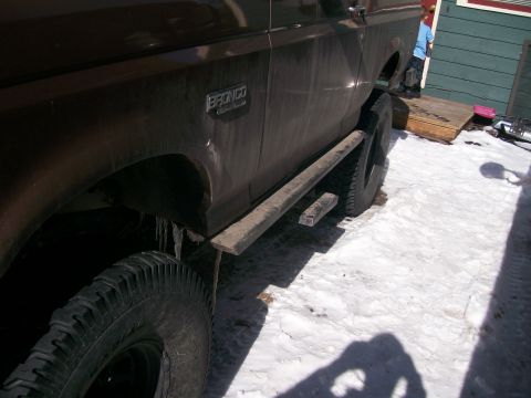 1993 Ford Bronco - Low side view along the driver's flank of a dirty dark brown Ford Bronco parked on packed snow, with icicles hanging from the fender lip an…