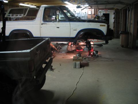 1985 Ford Bronco - A mechanic works on the undercarriage of a white and black 1985 Ford Bronco, which is raised on lifts in a garage setting.