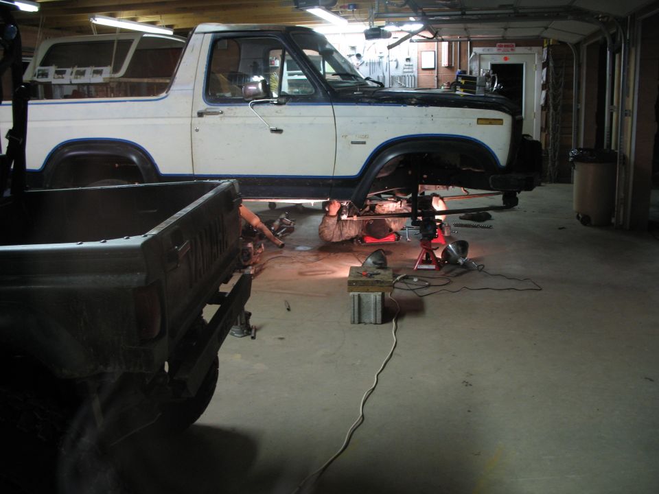 White and black 1985 Ford Bronco on lifts with a mechanic underneath