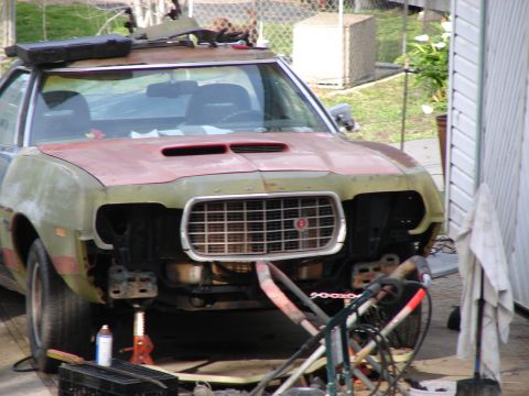 1973 Ford Ranchero - Front view of a stripped 1970s Ford project car sitting on a jack stand in a carport, with headlights, bumper, and front valance removed to…