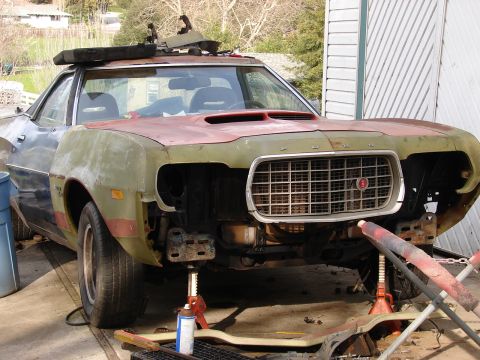 1973 Ford Ranchero - A 1970s Ford Ranchero-style car-pickup mid-restoration in a driveway, front bumper and headlights removed to expose the radiator support an…