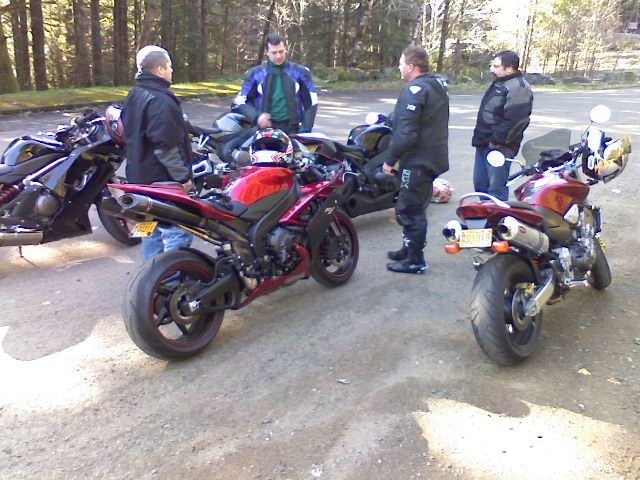 Four guys in motorcycle gear stand around a few sportbikes parked on a dirt lot