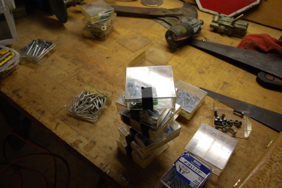 Workbench surface with stacks of clear plastic containers holding screws, nails, and bolts. A handsaw and other tools are visible in the background