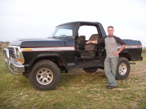 1978 Ford Bronco - Black full-size Ford Bronco with the top removed, tube door bars and cloth bucket seats visible, parked on desert grass while a man in a gr…