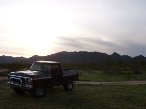 1978 Ford Bronco - Dark full-size Ford Bronco with its top removed parked on desert grass at sunset, backlit by the sun setting over low rocky mountains. The…