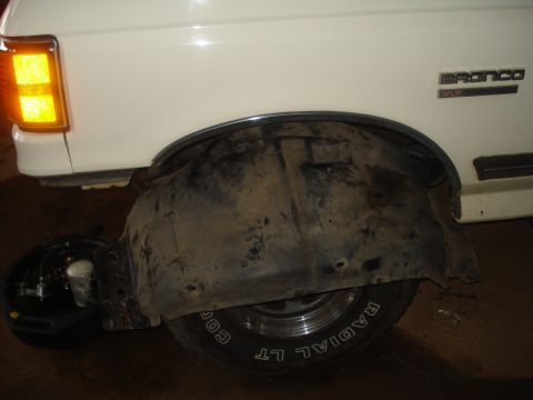 1991 Ford Bronco - The rear passenger side of a white Ford Bronco is shown, with the wheel well liner removed and a black drain pan with fluid underneath. A '…