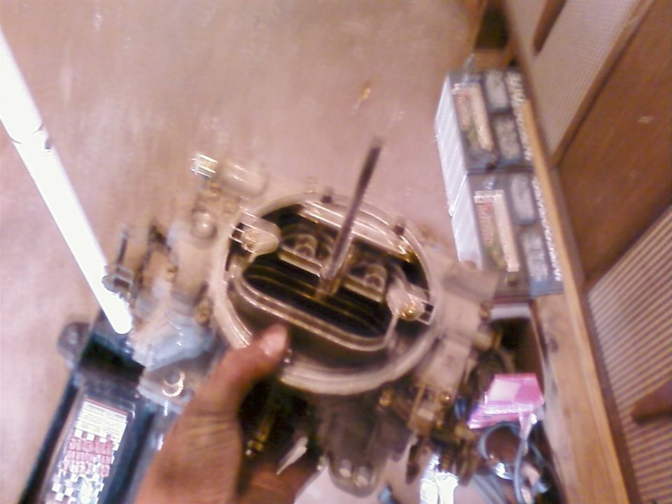 Close-up of a carburetor held by a hand, with automotive batteries in the background.