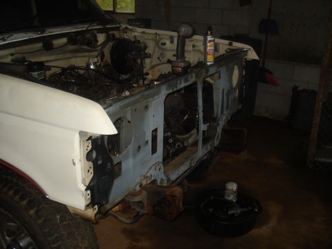 1991 Ford Bronco - The front end of a white 1991 Ford Bronco is shown disassembled in a garage. The engine bay is exposed, with various tools and parts scatte…
