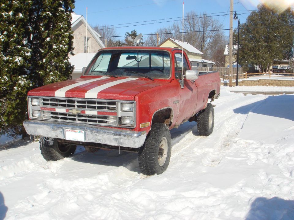 Lifted red 1985 Chevrolet C/K pickup truck with white hood stripes in the snow.
