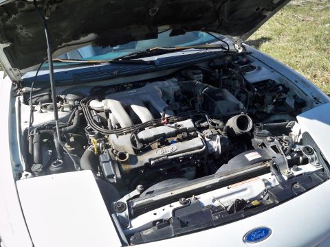 1995 Ford Probe - The open hood of a white 1995 Ford Probe reveals the engine bay with the V6 engine visible. The car is parked outdoors on a grassy area.