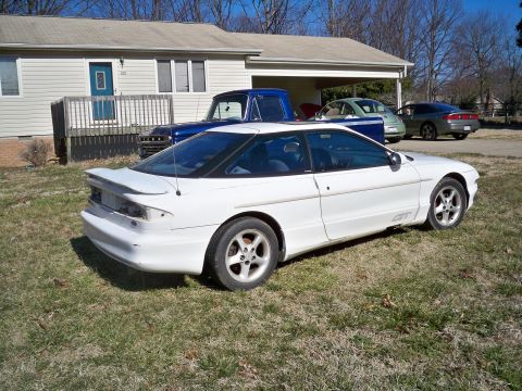 1995 Ford Probe - A white 1995 Ford Probe GT is parked on a grassy lawn in front of a house. A blue Ford pickup truck is parked behind it, partially covered…