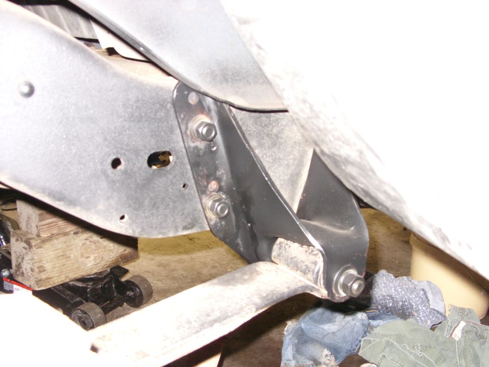 Underside of vehicle frame with black bracket, jack, and wood blocks.