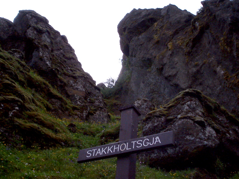 Brown 'STAKKHOLTSGJÁ' trail sign below mossy rocky cliffs