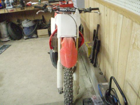 2005 Honda CR125R - A red and white dirt bike is parked in a garage, facing forward with its front wheel and handlebars clearly visible.