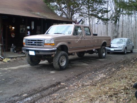 1979 Ford Bronco - Tan Ford F-Series pickup truck with large tires parked outside a cabin, a silver sedan is visible in the background.