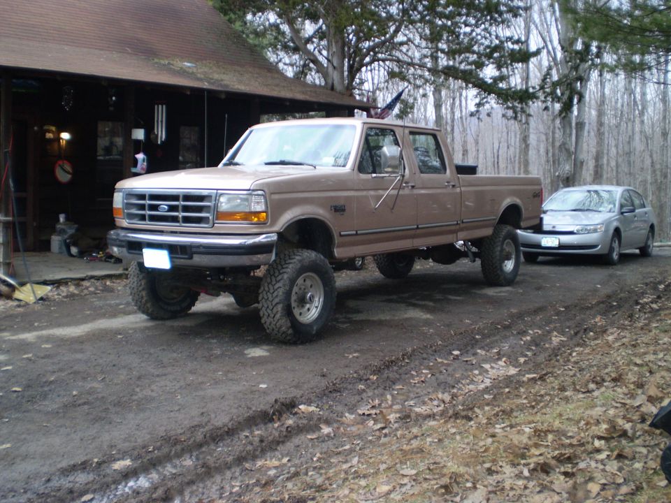 Lifted tan Ford crew cab pickup on mud tires parked on a dirt drive by a cabin