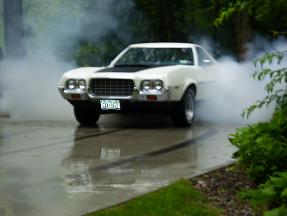 1972 Ford Ranchero - Small, low-resolution shot of a white Ford Ranchero doing a burnout on a wet concrete driveway, front three-quarter view with quad headligh…