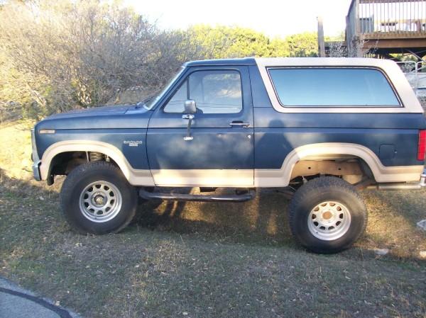 Blue 1986 Ford Bronco with tan trim and lifted suspension parked on grass.